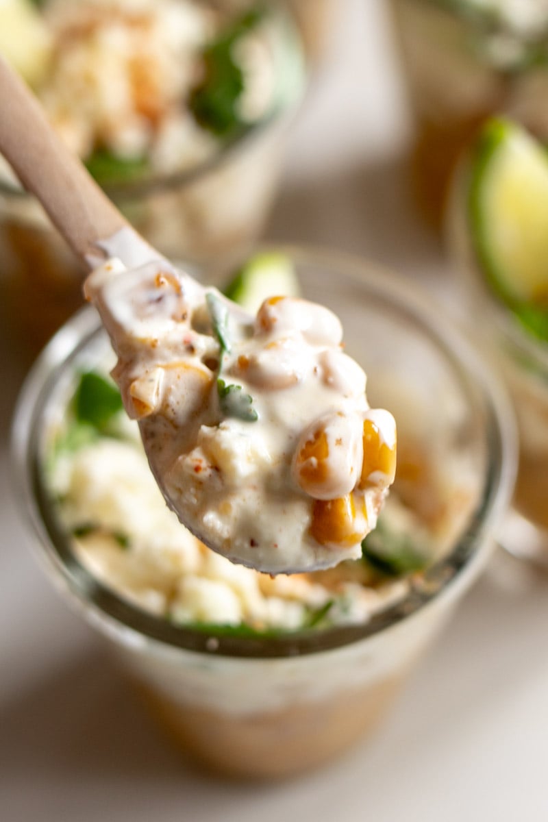 a wooden spoon holding a scoop of elote from a glass jar. 