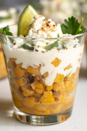 Elote in a glass cup with sour cream and lime on top.
