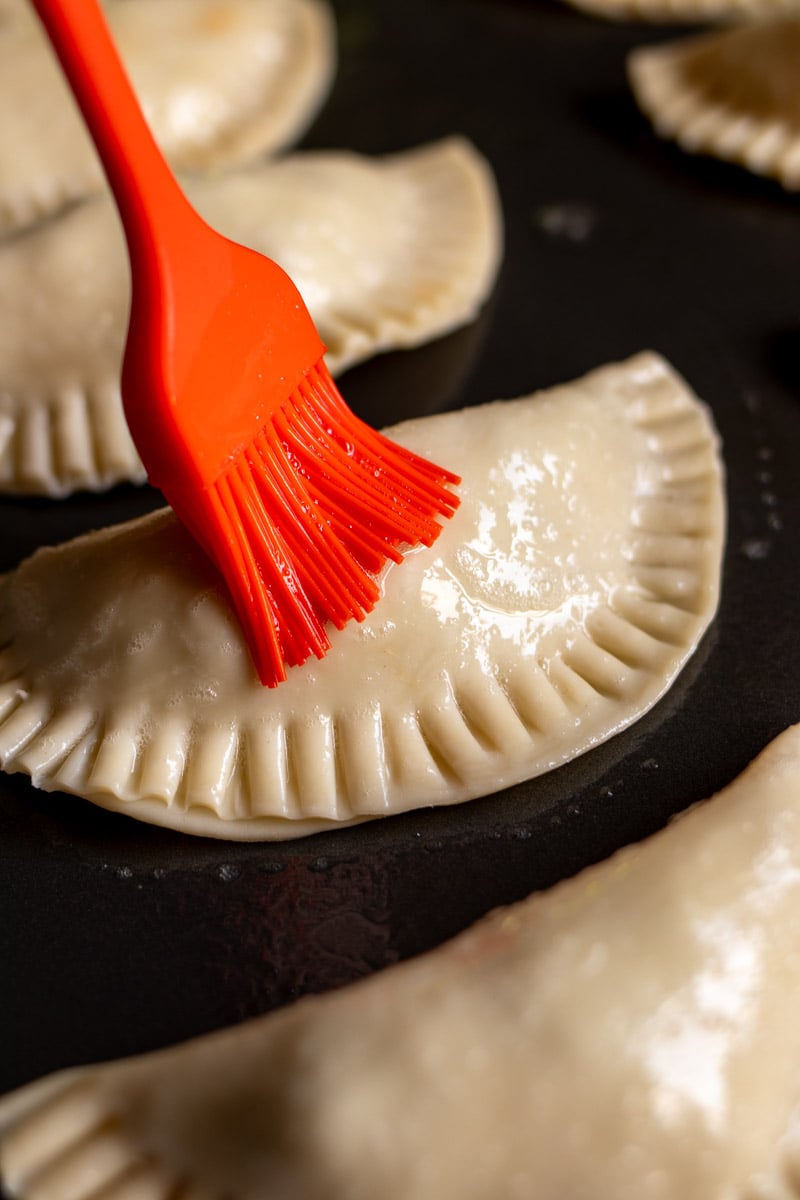 empanadas getting brushed with egg wash on a baking sheet. 