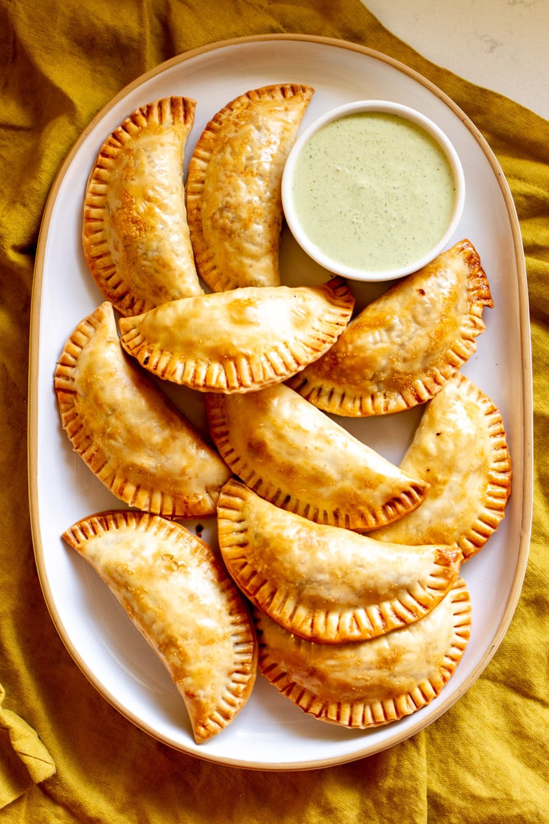 a white platter filled with beef empanads and a small bowl of dipping sauce. 