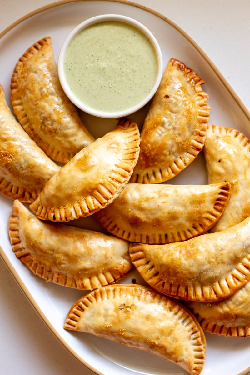 a white platter filled with golden brown baked empanadas that are filled with ground beef. 