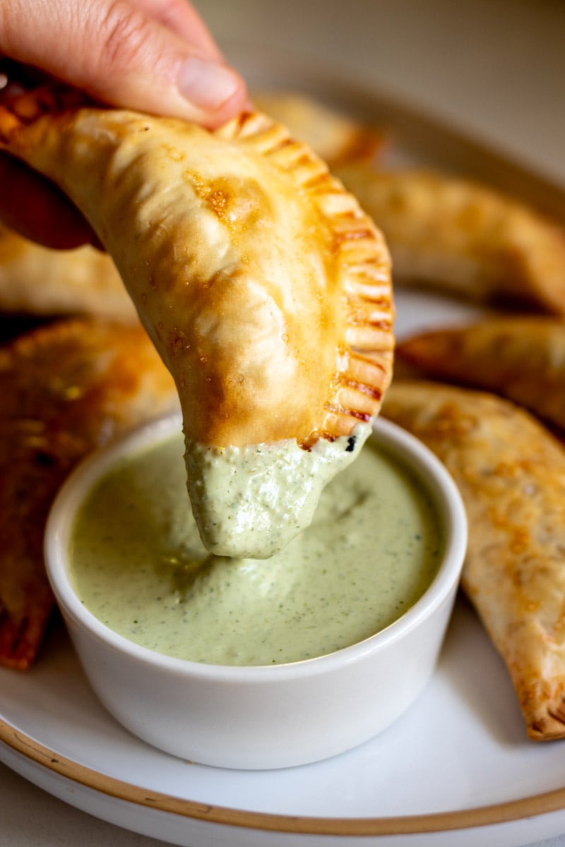 a hand dunking a beef empanada into a creamy green sauce. 