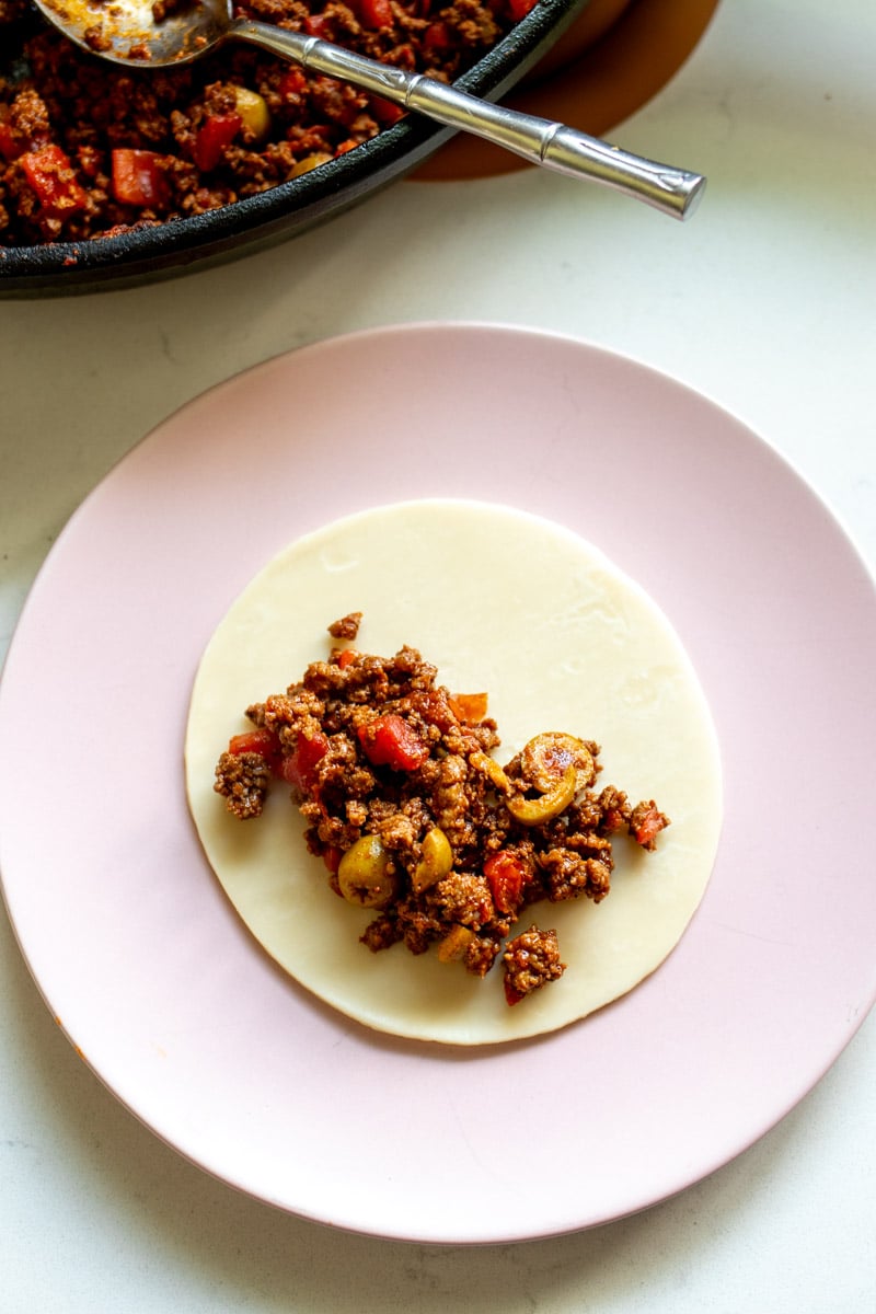 empanada dough topped with a tablespoon of meat filling on a pink plate. 