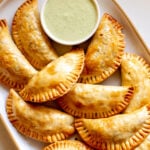 a white plate filled with baked empanadas and green sauce.