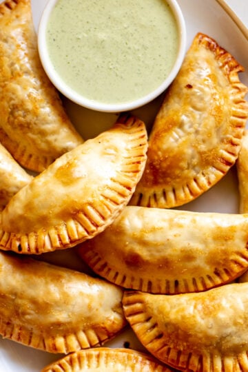 a white plate filled with baked empanadas and green sauce.
