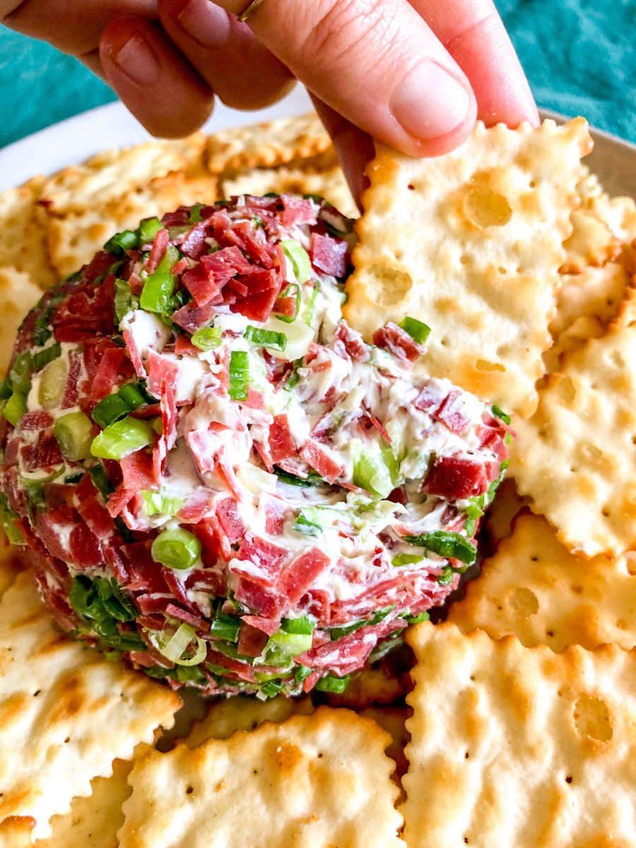 a hand holding a cracker scooping up some dried beef cheese ball.