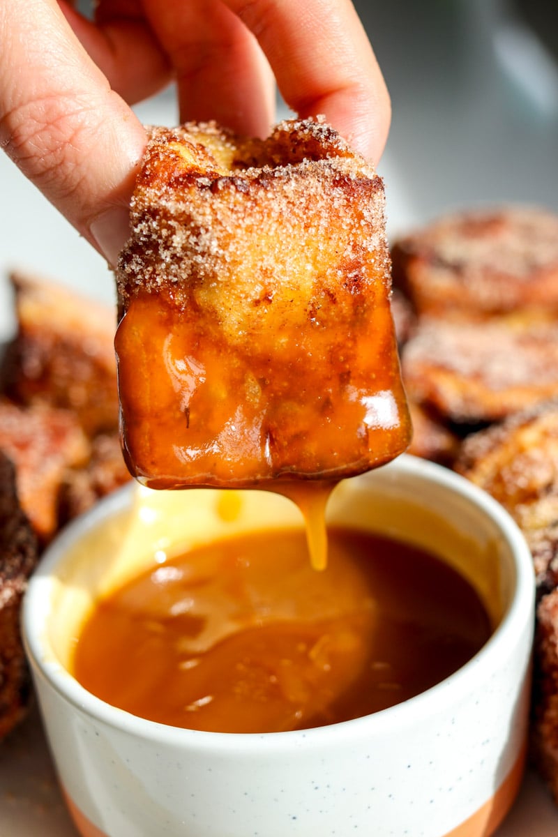 a hand holding a piece of hawaiian roll french toast dipping it into salted caramel.