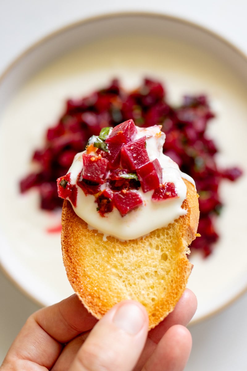 a hand holding a piece of toasted baguette that's been dipped into the hot honey whipped feta and pickled beet mixture.