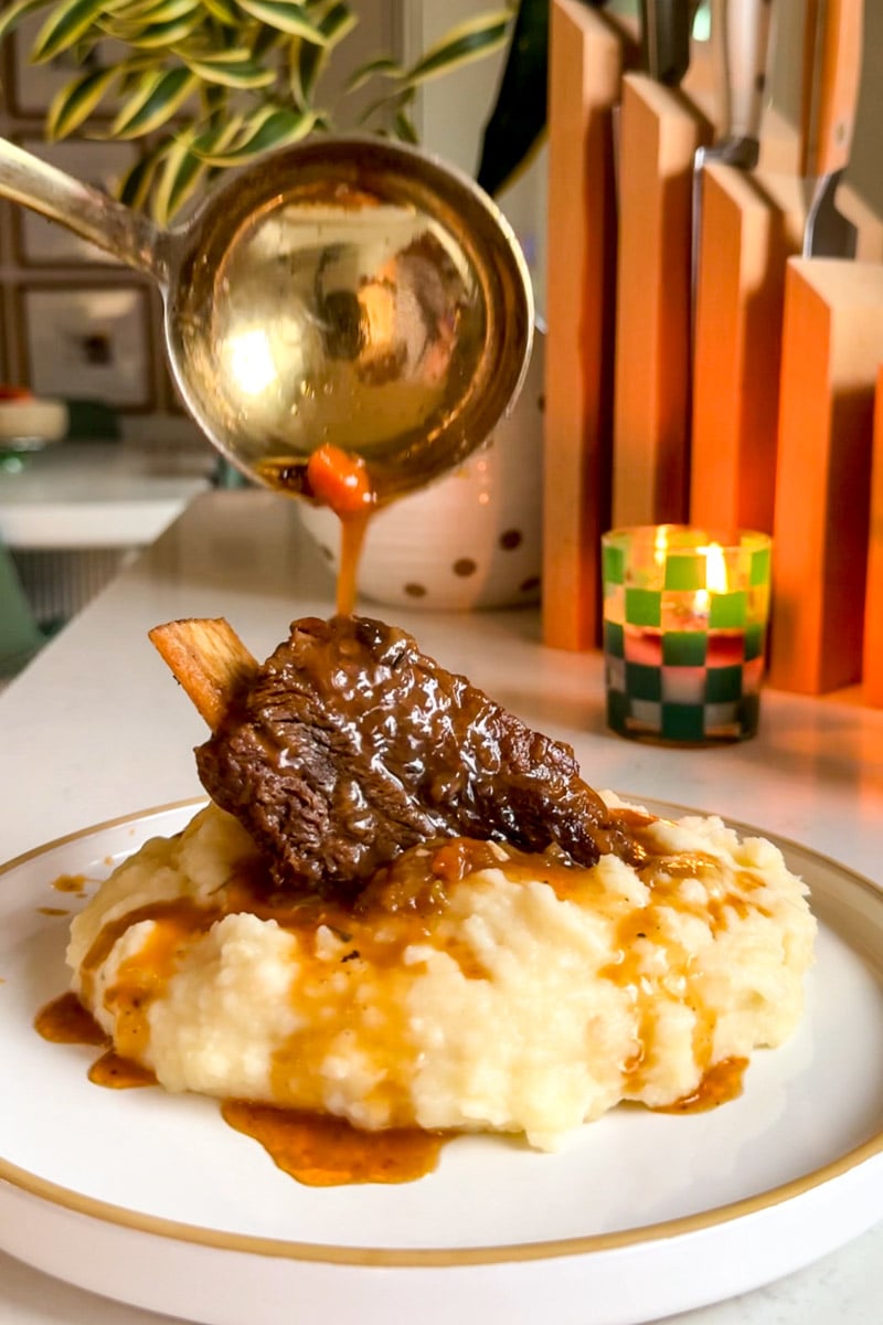 a chuck short rib on a white plate with a golden spoon ladeling gravy over it. 