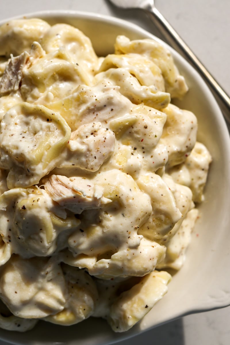 the right side of a serving bowl full of chicken tortellini alfredo with a fork in the background.