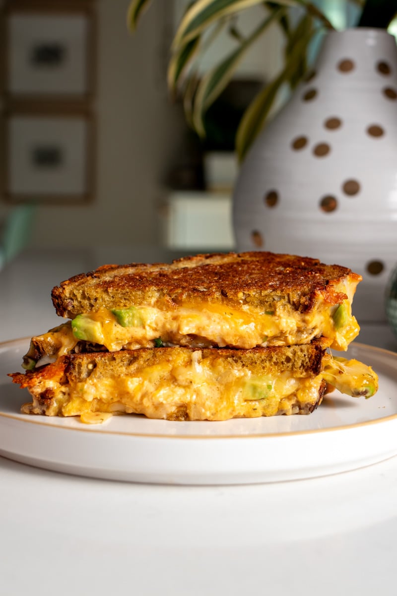 two halves of a buffalo chicken dip grilled cheese with sliced avocado on a white plate.