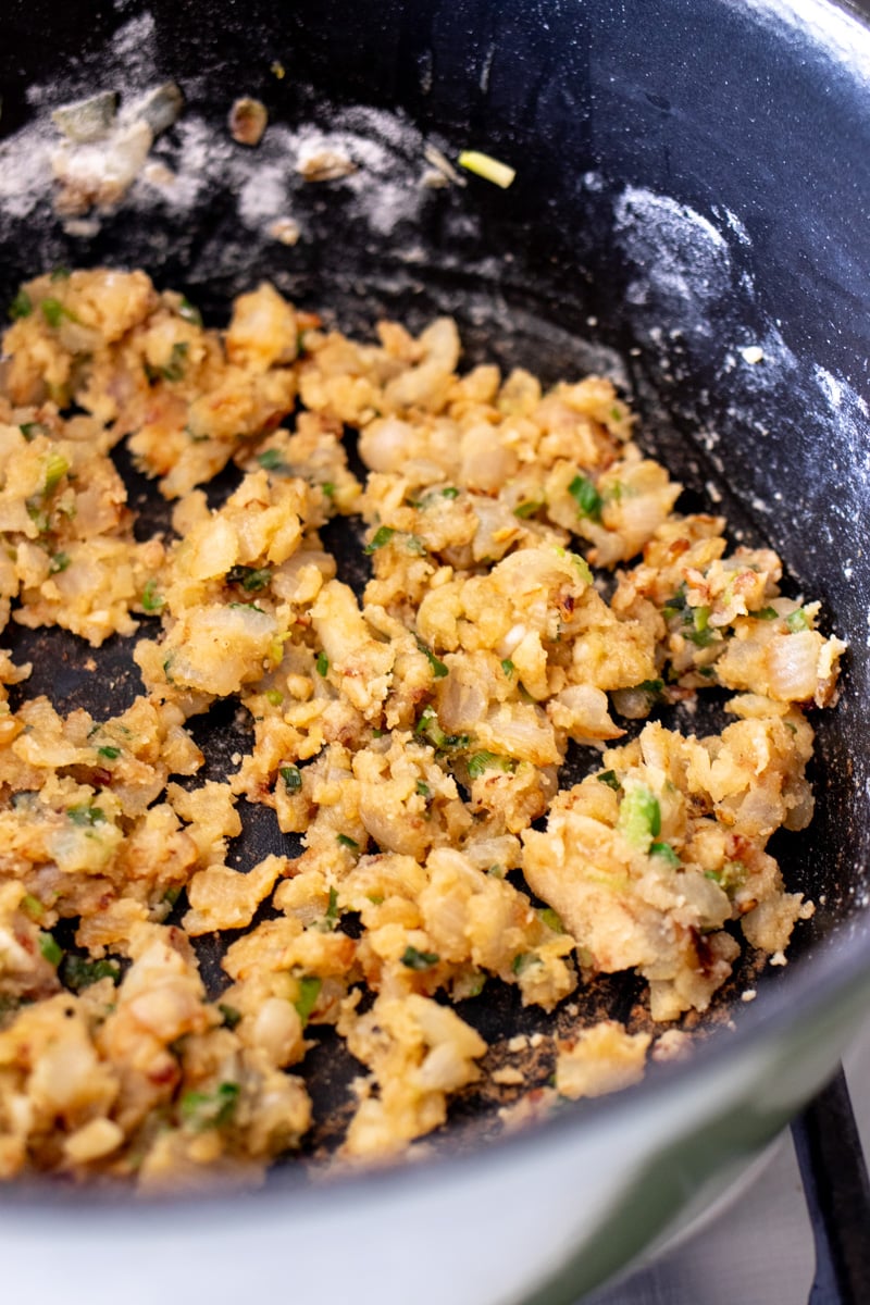 flour, onions, garlic and scallions being cooked in a large black pot to make a roux. 