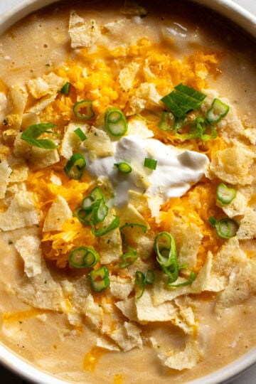 a white bowl filled with cheesy potato soup that's topped with cheddar, green onions and sour cream and onion potato chips.