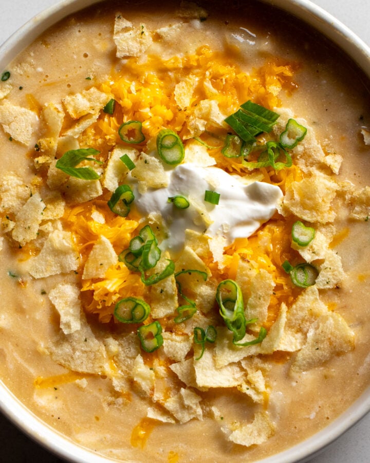 a white bowl filled with cheesy potato soup that's topped with cheddar, green onions and sour cream and onion potato chips.