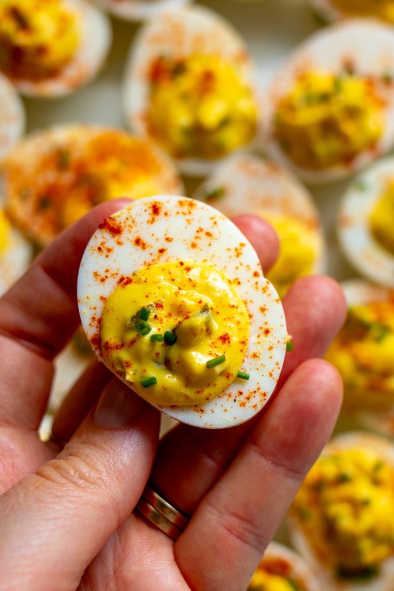 a hand holding a single deviled egg over a platter of them.