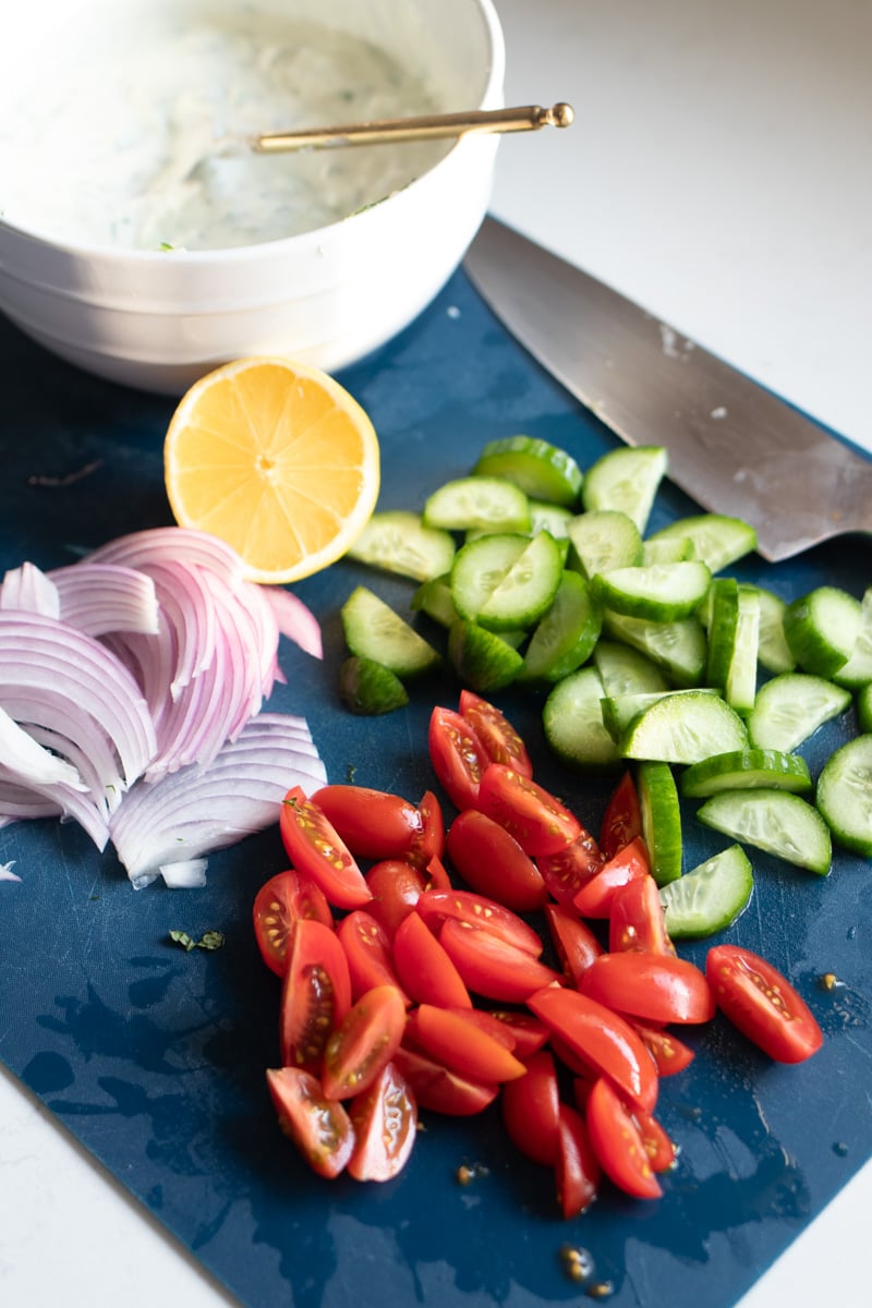 chopped up colorful cucumbers, tomatoes and red onion with a wedge of lemon and a bowl of tzatziki on a blue cutting board.