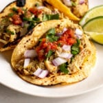 a close up of 3 instant pot chicken tacos topped with cilantro, onion and salsa with limes in the background.