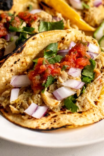 a close up of 3 instant pot chicken tacos topped with cilantro, onion and salsa with limes in the background.