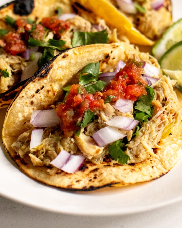 a close up of 3 instant pot chicken tacos topped with cilantro, onion and salsa with limes in the background.