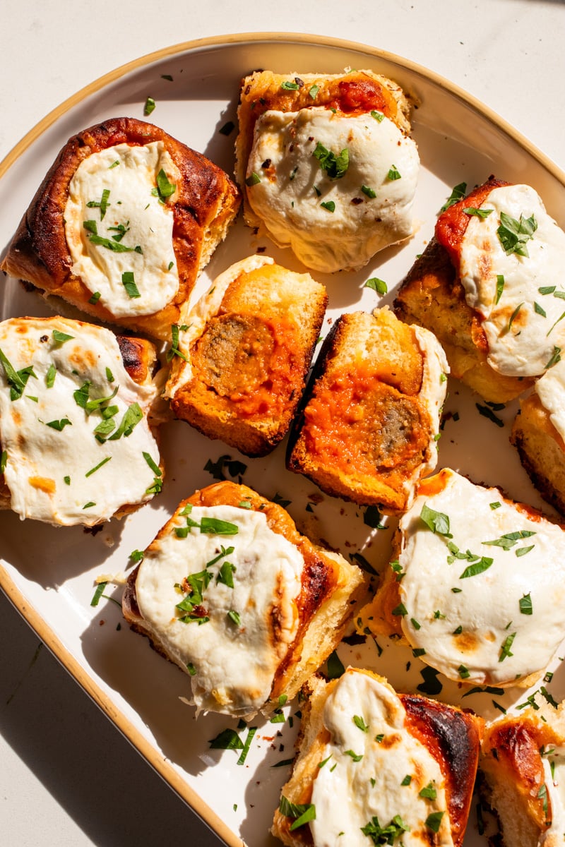 a white plate full of cheesy meatball sliders with one split in half so you can see the meatball and sauce.