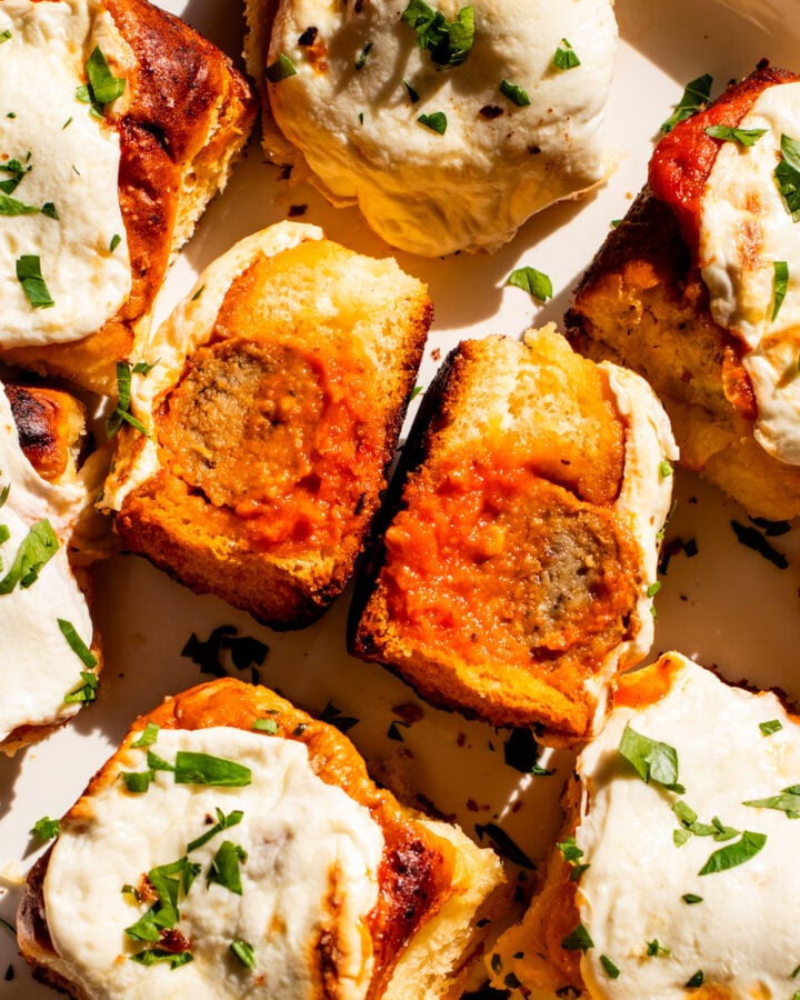 a white plate full of cheesy meatball sliders with one split in half so you can see the meatball and sauce.