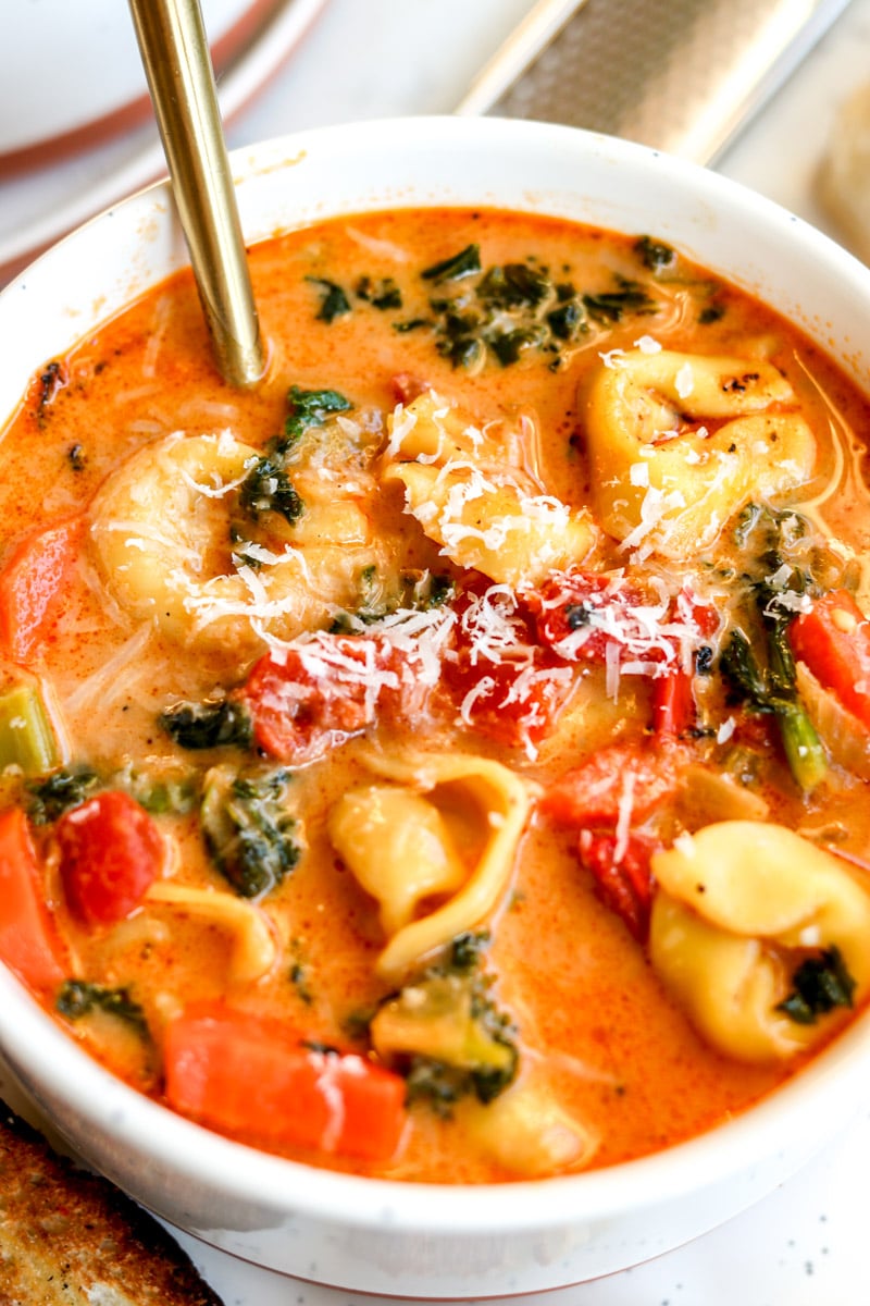 an overhead view of a big bowl of cheesy vegetarian tortellini soup with a golden spoon in it. 