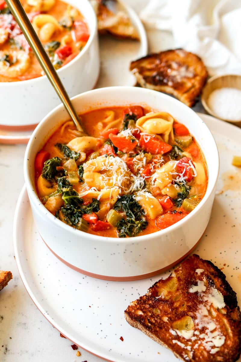 a white bowl filled with orange soup that's got cheese stuffed pasta and fresh vegetables.