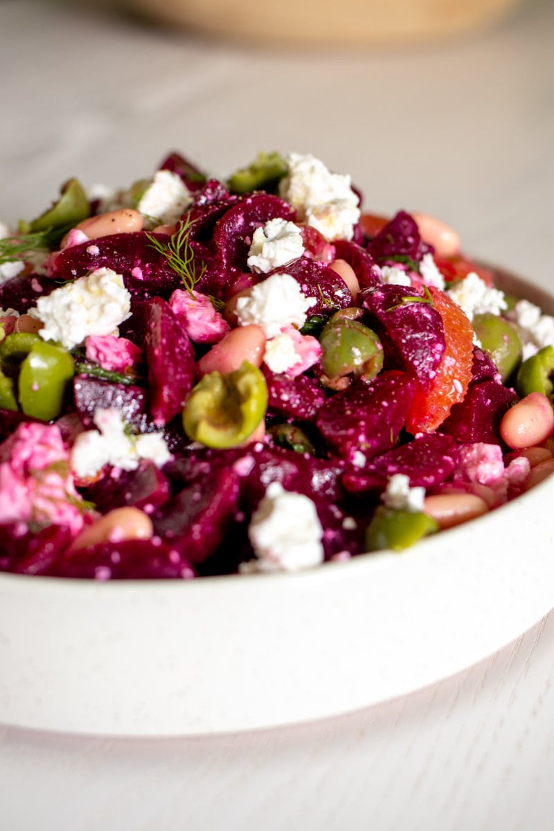 a white bowl filled with beet and feta salad, fresh herbs, oranges and olives.