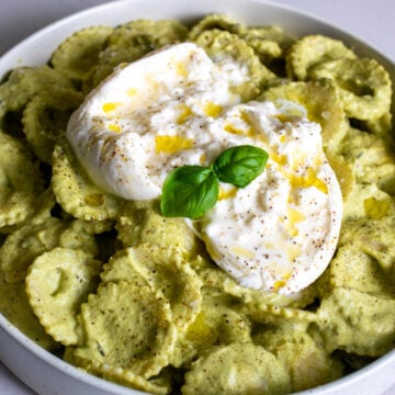 a white bowl of bright green broccoli pesto pasta with a ball of gooey burrata on top.