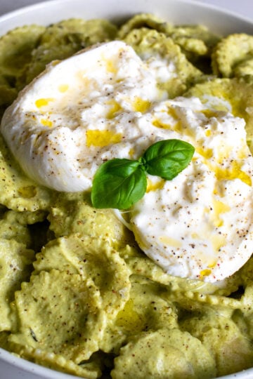 a white bowl of bright green broccoli pesto pasta with a ball of gooey burrata on top.