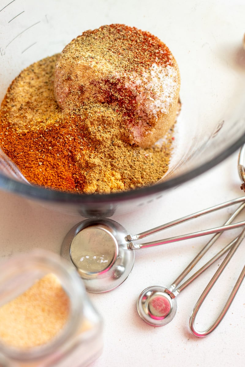different spices in a measuring cup before being stirred with measuring spoons in the foreground. 