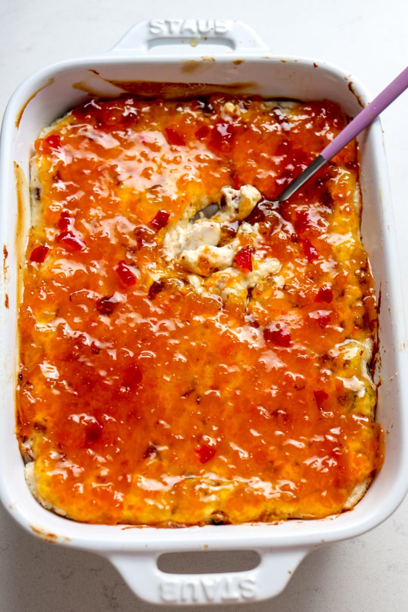 a white baking dish full of pepper jelly cheese dip with a spoon taking a dip out of it.