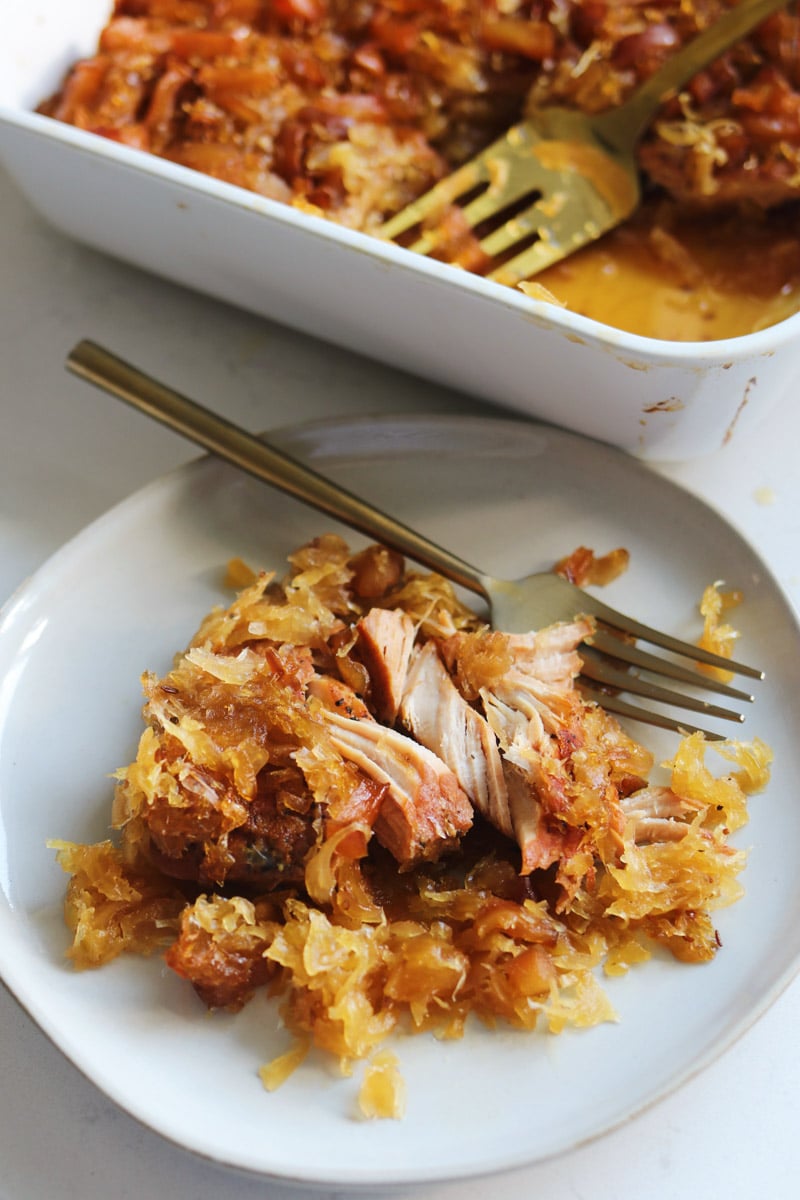 a white plate of pork and sauerkraut with a baking dish behind it.