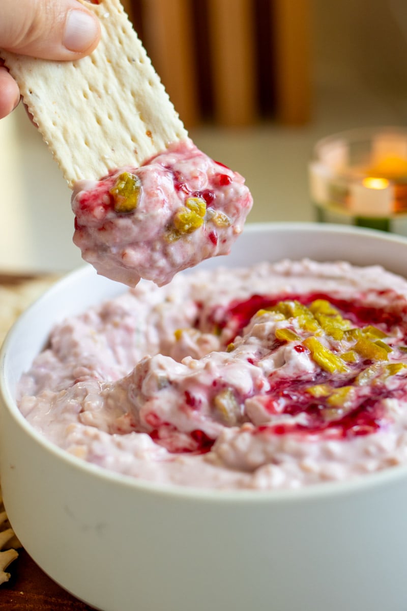 a cracker dipping into pink dip made with raspberry jam, creamy cheese and jalapenos.