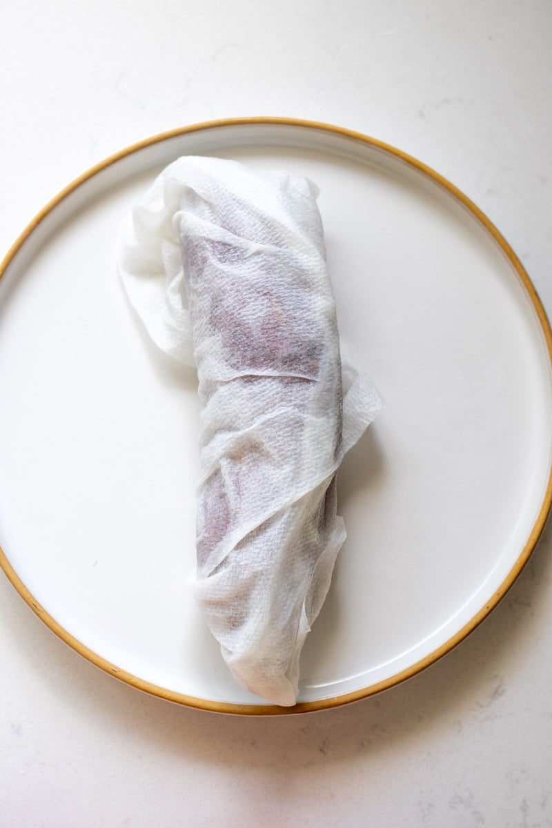 a sweet potato wrapped in a damp paper towel on a white plate.