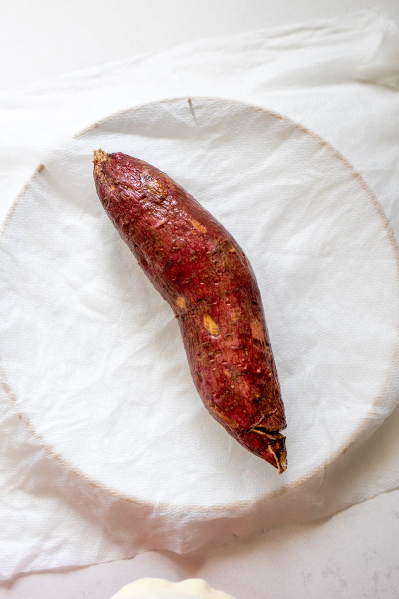 a washed and poked long sweet potato on a white plate with a paper towel near it.