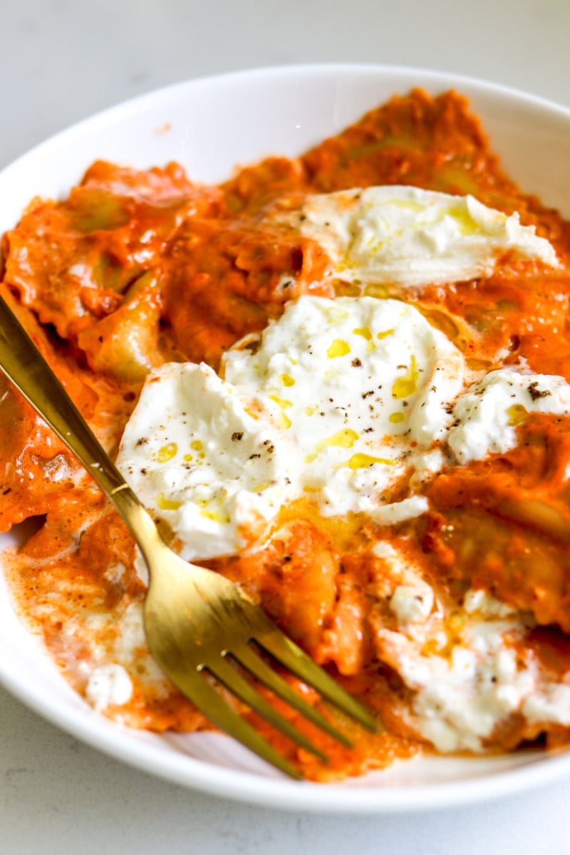 a white bowl filled with ravioli with tomato sauce and burrata on top. 