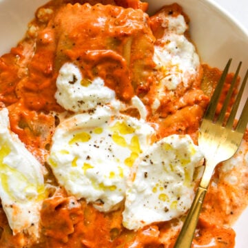 a white plate topped with orange red spicy tomato vodka sauce with ravioli underneath and burrata on top.