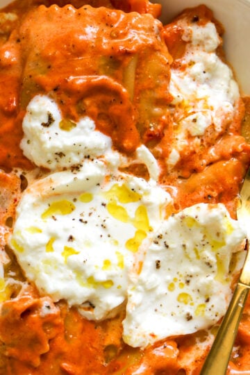 a white plate topped with orange red spicy tomato vodka sauce with ravioli underneath and burrata on top.