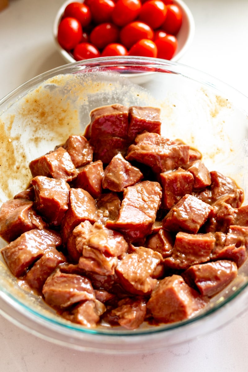 marinated petite sirloin steaks in a glass bowl with tomatoes behind it. 