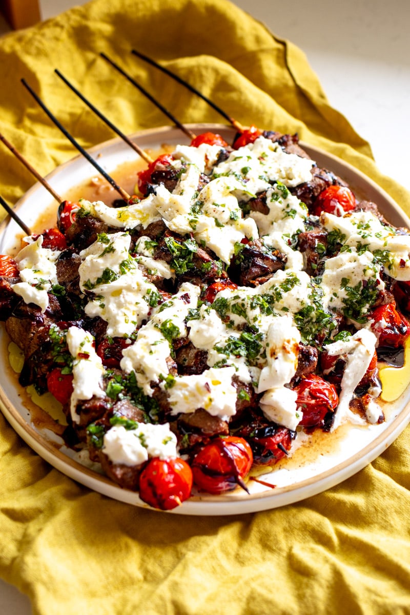 basalmic glazed steak skewers with tomatoes, burrata and garlic basil herb oil on top all on a white plate with a yellow napkin below.