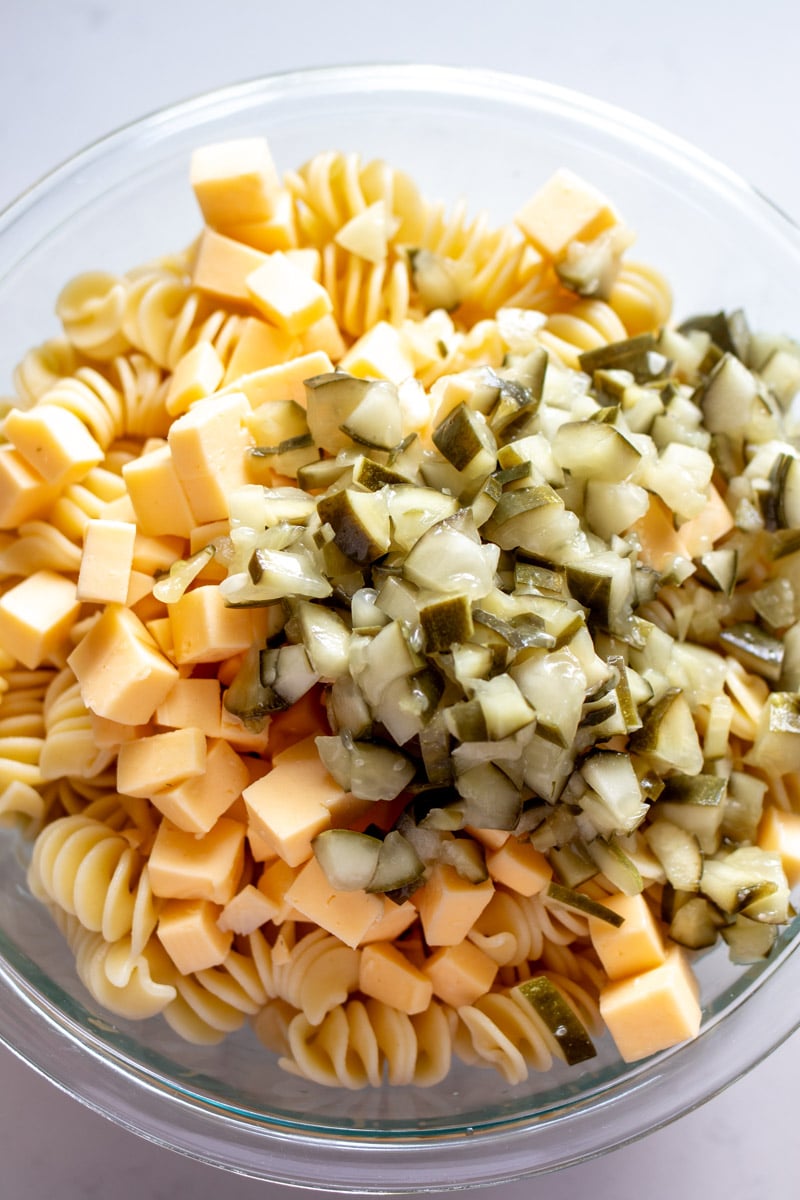 pasta,, pickles, cheese and dressing in a glass bowl before it's mixed.
