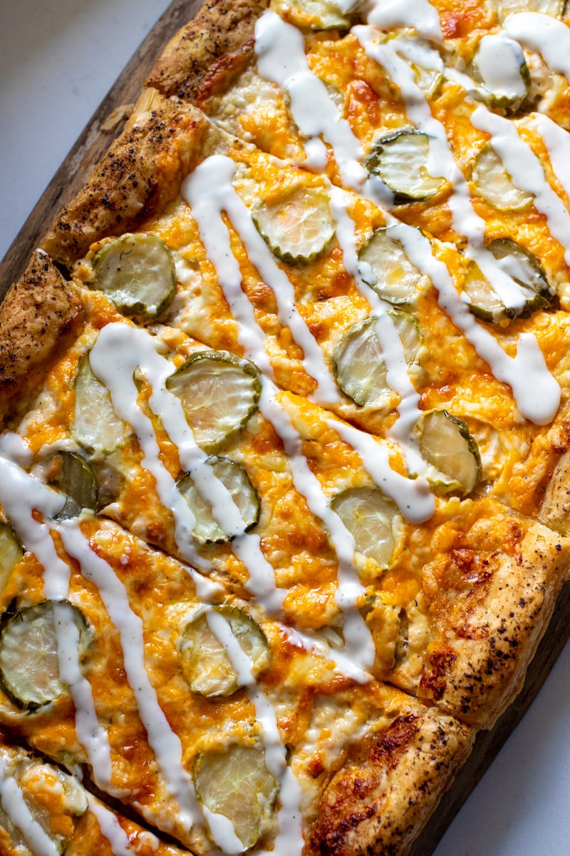 a dill pickle pizza drizzled with ranch dressing on a wooden cutting board.