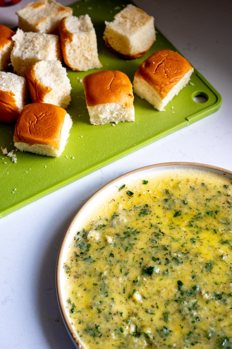 garlic butter mixture on a shallow plate with trimmed hawaiian rolls on a green cutting board in teh background.
