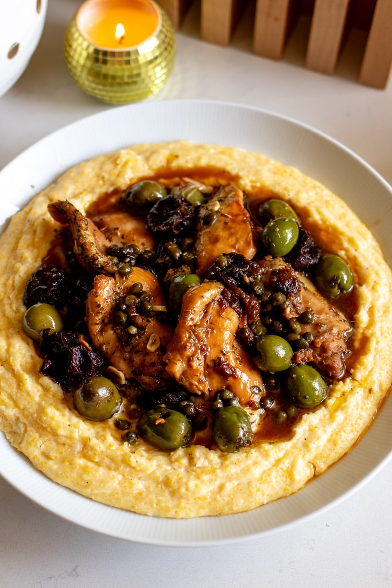 a big white bowl full of capers, olives prunes and tender chicken marbella over cheesy polenta.