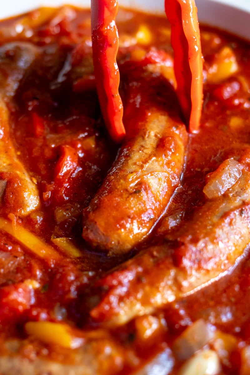 tongs holding a sausage over a crockpot full of sausage, peppers and onions.