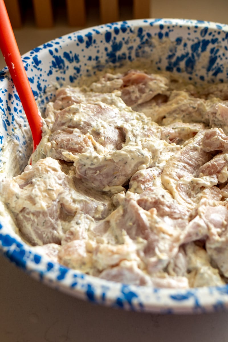chicken thighs marinating in greek yogurt, lemon, garlic and spices in a blue speckled bowl with an orange spatula.