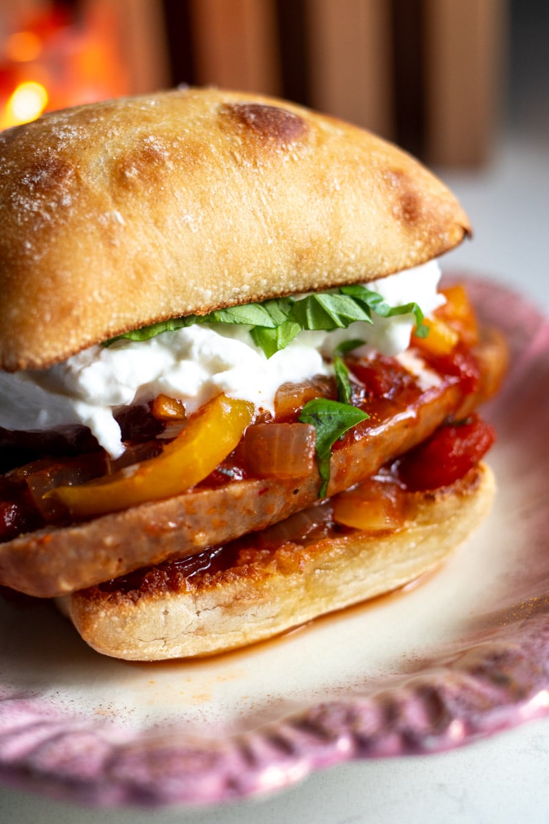 an italian sausage sandwiches loaded with peppers, onions, basil and burrata on a white plate.
