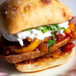 an italian sausage sandwiches loaded with peppers, onions, basil and burrata on a white plate.