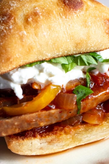 an italian sausage sandwiches loaded with peppers, onions, basil and burrata on a white plate.